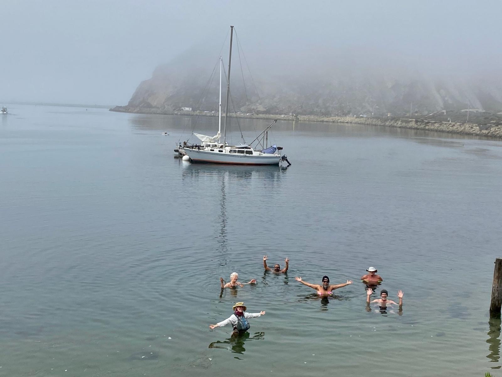 A group of odders with arms wide open in the water on a foggy morning, sailboat anchored behind them