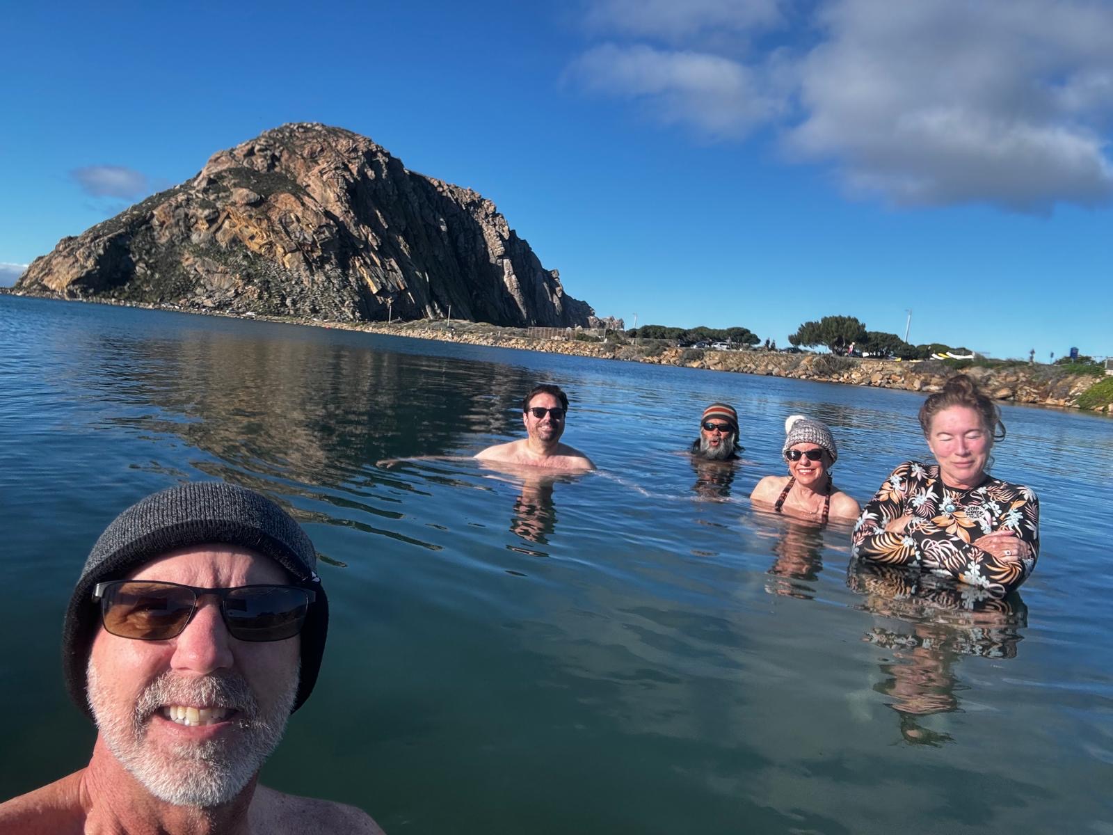 Five smiling club members shoulder to shoulder in the water with Morro Rock behind them